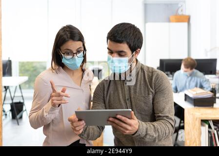 Uomo e donna ispanici che indossano maschere facciali protettive che chiacchierano mentre si guarda un tablet. Lavorare in ufficio durante il concetto pandemico di Coronavirus. Foto Stock