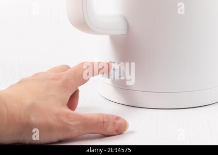 una mano femmina accende un bollitore elettrico bianco a. bollire l'acqua sullo sfondo di un tavolo di legno bianco Foto Stock