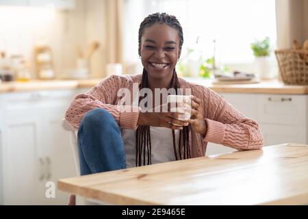 Giovane donna afroamericana allegra che si rilassa in una cucina accogliente, bevendo caffè Foto Stock