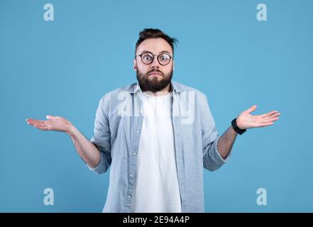 Chissà. Uomo confuso bearded in occhiali shrugging spalle su sfondo blu studio Foto Stock