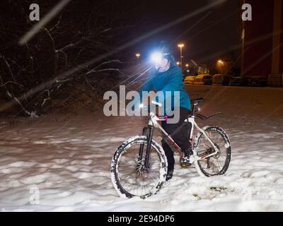 Ragazzo adolescente con bici in un parco innevato invernale. Il bambino tiene il manubrio e si siede su una bicicletta. Ragazzo con un caldo colore blu nero, casco e faro luminoso. Foto Stock