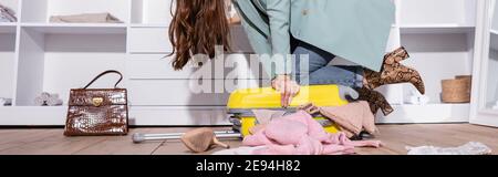 Vista ritagliata della donna chiusura valigia vicino vestiti sul pavimento, banner Foto Stock