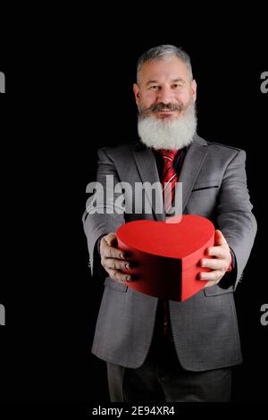 Mature uomo bearded abito grigio tiene rosso cuore forma scatola regalo, isolato sfondo nero Foto Stock