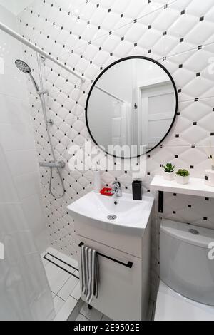 Luminoso bagno dal design moderno in un monolocale. Prevalentemente bianco con accenti neri. Uno specchio rotondo appeso sopra il lavandino sulla parete piastrellata Foto Stock