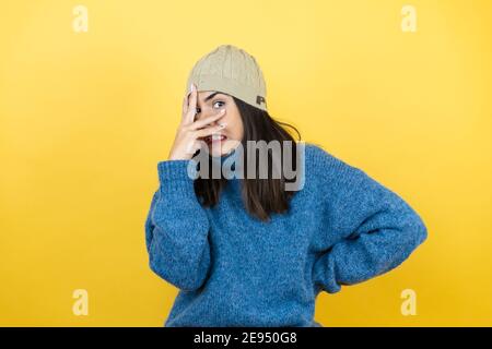 Giovane bella donna che indossa un pullover casual blu e cappello di lana che sbirciano in shock coprendo viso e occhi con la mano, guardando attraverso le dita con imbarra Foto Stock