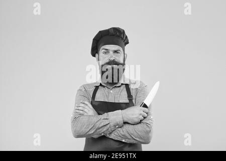 Provate la cucina raffinata. Il menu migliore offre un'uniforme rossa dello chef. Chef maschile tagliato con coltello. Hipster maturo con viso serio. Cuoco bearded ha attraversato le mani. Macellaio brutale in grembiule. Foto Stock