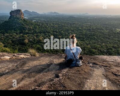 Giovane caucasica seduta sulla cima della roccia Pidurangala. Shot orizzontale. Da dietro. Sri Lanka, Asia. Foto Stock
