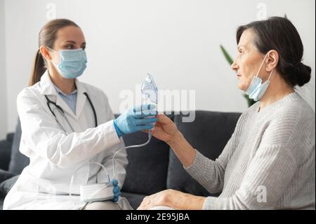 Una dottoressa, un'infermiera che indossa un abito medico e una maschera facciale, mette una maschera di ossigeno sul paziente anziano malato a casa durante la pandemia. Assistenza sanitaria e. Foto Stock