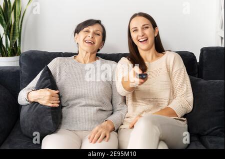 Una madre anziana e una figlia adulta si rilassano sul divano di casa, guardano la TV, cambiano i canali con il telecomando, sorridono e si godono il tempo Foto Stock