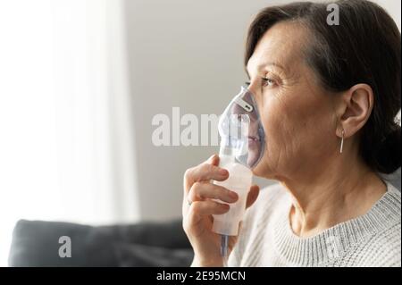 Vista laterale una donna anziana sta usando l'inalatore per il trattamento di flu e freddo seduto sul divano a casa. Concetto di sanità e medicina Foto Stock