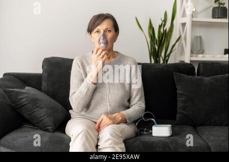 Donna anziana che usa l'inalatore per il trattamento dell'influenza e del freddo seduto sul divano a casa. Concetto di sanità e medicina Foto Stock