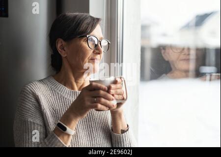 Primo piano ritratto della donna anziana anziana che indossa occhiali gode di caffè mattutino in cucina a casa. Una moderna signora pensante che sogna con una tazza Foto Stock