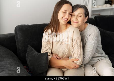 Una figlia adulta e una madre anziana si siedono sul comodo divano in abbraccia godendo il tempo insieme con gli occhi chiusi, a lungo non si sono visti l'un l'altro Foto Stock