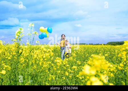 campo giallo, cielo blu, nuvole bianche. Corsa bambini carini con palloncini blu e gialli. Il concetto di libertà, vacanza. Cartolina estiva. Foto Stock
