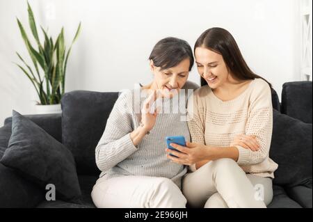 Una figlia adulta e una madre anziana che utilizzano lo smartphone per videoconferenze, videochiamate, due donne multigenerazionali sedute sul divano e guardate Foto Stock