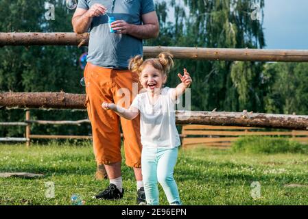 Un papà e sua figlia stanno facendo bolle nel Park.cheerful bella bambina corre e ride, prende bolle. Felice infanzia. Estate soleggiata Foto Stock
