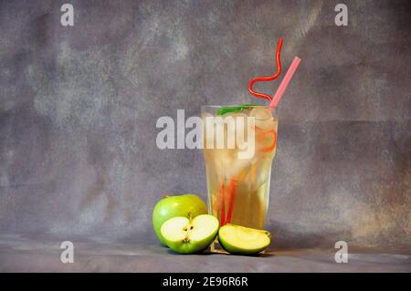 Un alto bicchiere di nettare di frutta con ghiaccio e tubuli, mele verdi mature accanto ad esso su sfondo grigio. Primo piano. Foto Stock