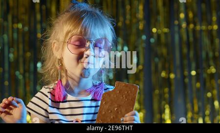 Primo piano ritratto di elegante bambino trendy al club discoteca party cyberpunk mangiare grande cioccolato bar caramelle dolci. Giovane giocosa ragazza bionda con colorate luci blu al neon su sfondo lucido Foto Stock