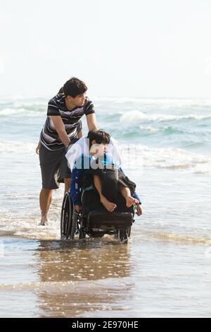 Fratello maggiore che spinge il ragazzino disabile in sedia a rotelle sulla spiaggia attraverso l'acqua dell'oceano lungo la riva Foto Stock