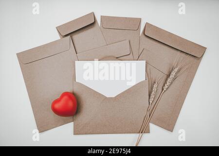 La carta bianca bianca bianca è posta sulla busta di carta marrone aperta con il cuore rosso e il fiore asciutto di Barley su sfondo bianco. Concetto di San Valentino. Foto Stock