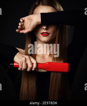 Giovane bella donna con lunghi capelli setosi in abiti neri utilizza una piastra per capelli rossa e copre gli occhi con l'altro mano Foto Stock