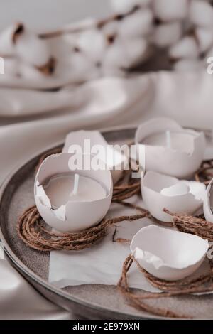 Decorazioni pasquali alla moda. Uova organiche cracked su piastra. Candela in guscio bianco delle uova. Piuma d'uccello. Spazio di copia. Biglietto d'auguri. Colori pastello. Fai da te di Pasqua. Fai da te. Foto Stock