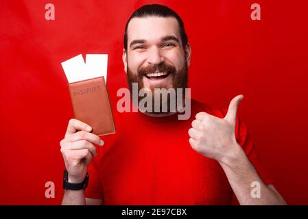 Un uomo felice sta tenendo un passaporto con due biglietti in esso e mostrando un pollice in su. Foto Stock