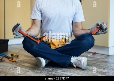 Colpo corto di giovane handyman che tiene un martello e una chiave regolabile nelle sue mani mentre medita, seduto in posa di loto sul pavimento all'interno Foto Stock