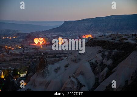 Le luci notturne della città di Goreme e i mongolfiere si illuminano mattina presto, mentre si gonfia tra le rocce di paesaggio di montagna in Cappadocia Foto Stock