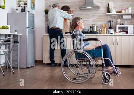 Donna anziana paralizzata in sedia a rotelle sentendosi infelice seduto nel mezzo della cucina mentre il marito disimballare la borsa della spesa. Vecchia signora handicappata dopo lesione e riabilitazione, depresso non valido pieno di dolore Foto Stock