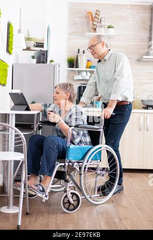 Paralysied moglie anziana in sedia a rotelle che lavora al computer tablet, mostrando a marito il suo progetto seduto in cucina. Persone anziane disabili che usano la moderna tecnologia internet online. Foto Stock