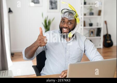 Divertente ritratto di gioioso dipendente africano di ufficio maschile seduto a. la scrivania con un computer portatile che indossa una maschera da snorkeling e guarda alla macchina fotografica con sorriso allegro Foto Stock