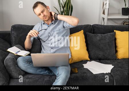Il freelancer overworked prende una rottura nel mezzo di una giornata estenuante. Un giovane stanco con un computer portatile si siede sul divano e tiene la nuca Foto Stock