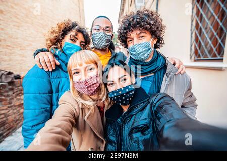 Amici multirazziali coperti da maschera facciale che prende selfie indossando l'inverno Vestiti - nuovo concetto di amicizia normale con i giovani millenari divertirsi Foto Stock