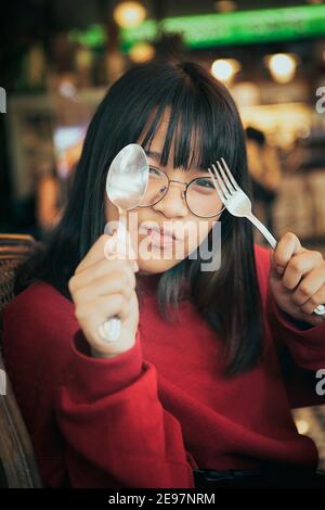 faccia divertente dell'adolescente asiatico solleva forchetta e cucchiaio sala da pranzo Foto Stock