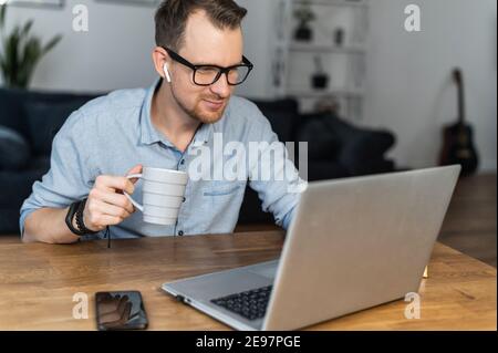 Un ragazzo intelligente che indossa occhiali e una camicia casual elegante si siede alla scrivania con una tazza di caffè e utilizza un computer portatile per lavorare a distanza. Si sviluppa un giovane Foto Stock