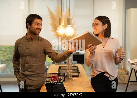 Felice uomo ispanico dando una cartella alla sua donna sorridente collaboratore mentre si guarda l'un l'altro. Buon concetto di ambiente di lavoro. Foto Stock