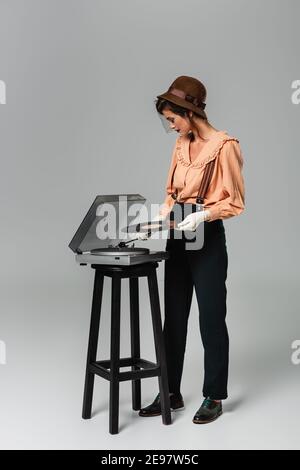 vista completa della bella donna in cappello ed elegante abiti retrò che mettono il disco in vinile sul lettore di dischi in grigio Foto Stock