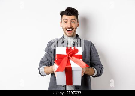 Sorpreso giovane uomo sorridente eccitato, tenendo grande scatola regalo sulle feste di giorno di valentines, ricevere sorpresa presente, in piedi stupito su sfondo bianco Foto Stock