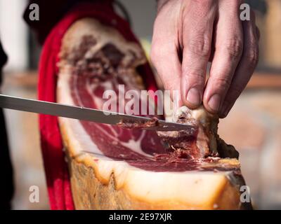 Vista ad angolo basso dell'uomo che taglia una fetta di jamon spagnolo serrano secco su un tavolo di legno. Foto Stock
