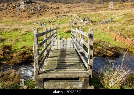Passerella in legno che attraversa un ruscello con camminatori lontani sul sentiero nel Parco Nazionale di Snowdonia. Bethania, Beddgelert, Gwynedd, Galles, Regno Unito, Gran Bretagna Foto Stock