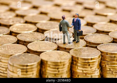 Primo piano di imprenditori maschili in miniatura in piedi su pila di euro centesimi e mani agitanti mentre si raggiunge un accordo su deal Foto Stock