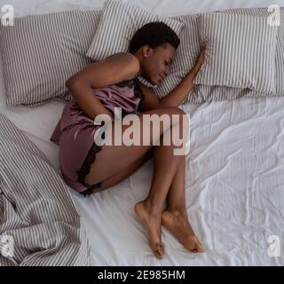 Infelice stressata millenaria donna afroamericana sdraiata a letto raddoppiata con dolorosa sensazione di stomaco, malsana Foto Stock