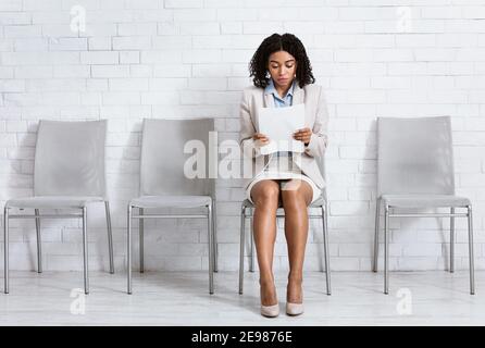 Giovane donna nera in abbigliamento formale si sente preoccupata prima del suo colloquio di lavoro, in attesa in sala aziendale, rileggendo il suo CV Foto Stock