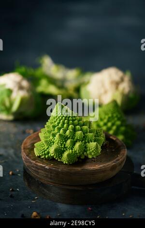 primo piano di una testa di romanesco broccoli bambino su un piatto di legno, posto su una superficie di pietra scura accanto ad un altro bambino romanesco broccoli testa e alcuni Foto Stock