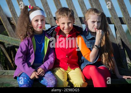 Bambini piccoli divertenti: Le ragazze con il trucco luminoso vestito nello stile degli anni novanta e un ragazzo in una camicia rossa sono seduti su una panchina da una recinzione inclinata Foto Stock