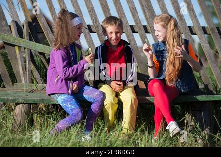 Bambini piccoli divertenti: Le ragazze con il trucco luminoso vestito nello stile degli anni novanta e un ragazzo in una camicia rossa sono seduti su una panchina da una recinzione inclinata Foto Stock