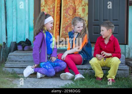 Bambini piccoli divertenti: Le ragazze con il trucco luminoso vestito nello stile degli anni novanta e un ragazzo in una camicia rossa sono seduti sul portico del villaggio del Foto Stock