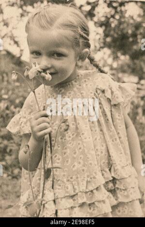 Lettonia - CIRCA 1930: Ritratto di ragazza che gioca con il fiore in giardino. Fotografia d'epoca Art Deco colorata d'epoca Foto Stock