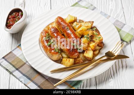 bangers e patate arrostite cosparse di timo fresco su un piatto bianco con sugo di cipolla in una brocca su un tavolo in legno testurizzato bianco Foto Stock